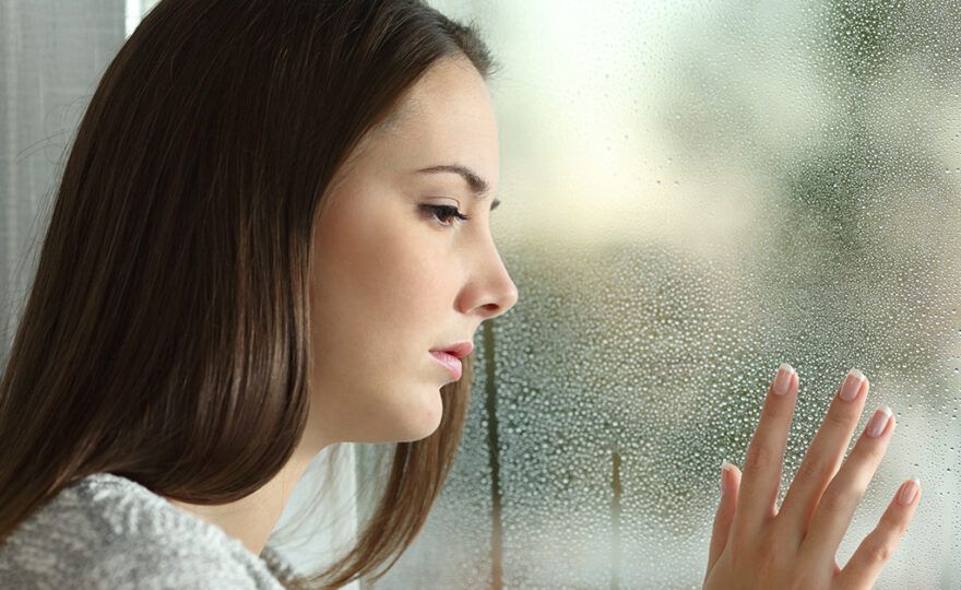 Sad woman looking the rain falling through a window at home