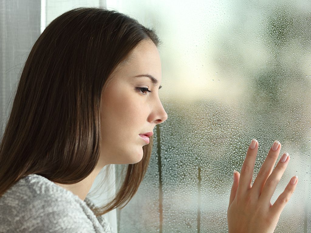 Sad woman looking the rain falling through a window at home